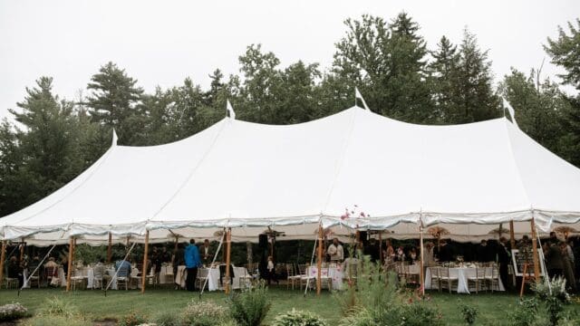 Large white outdoor tent with people and tables set up inside on a grassy area, surrounded by trees and plants.