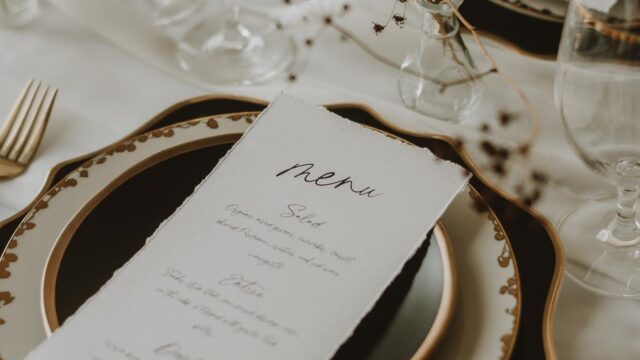 Elegant table setting with stacked plates, a detailed menu, and glassware, featuring gold accents and minimalist floral decorations.