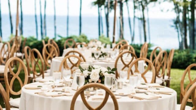 Outdoor event setup with round tables covered in white tablecloths and wooden chairs under a large tent, overlooking a scenic view of trees and water.