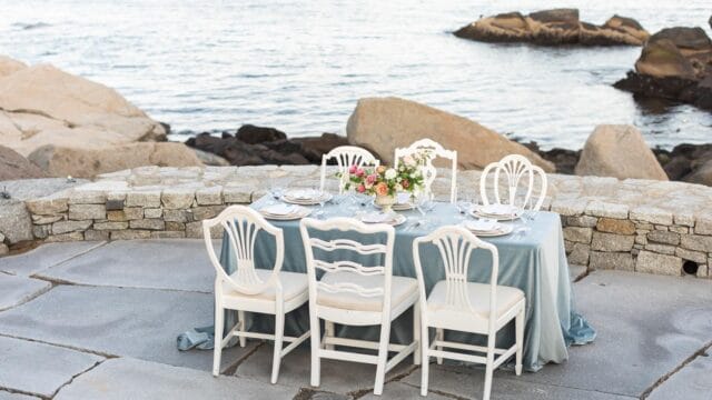 Elegant table setting with a blue tablecloth and white chairs on a stone patio by the sea, featuring a floral centerpiece and tableware.