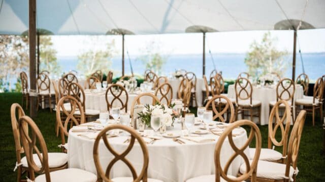 Outdoor wedding reception with round tables, white tablecloths, elegant chairs, and floral centerpieces under a tent, trees, and water in the background.