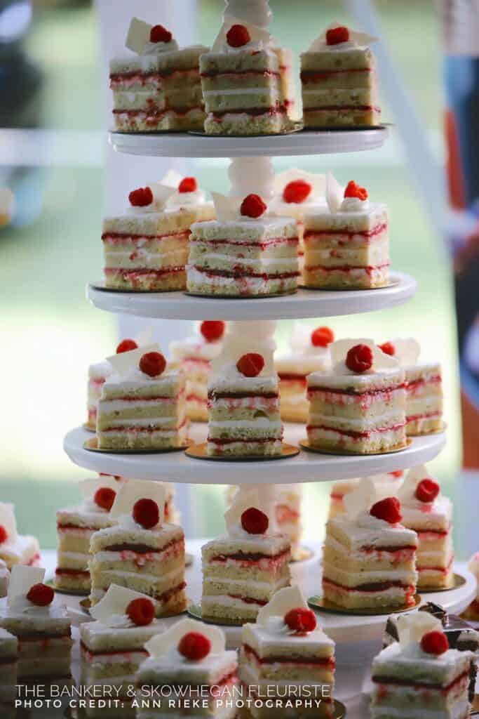 A tiered display of frosted square cakes with raspberries on top and white chocolate pieces is arranged on circular plates.