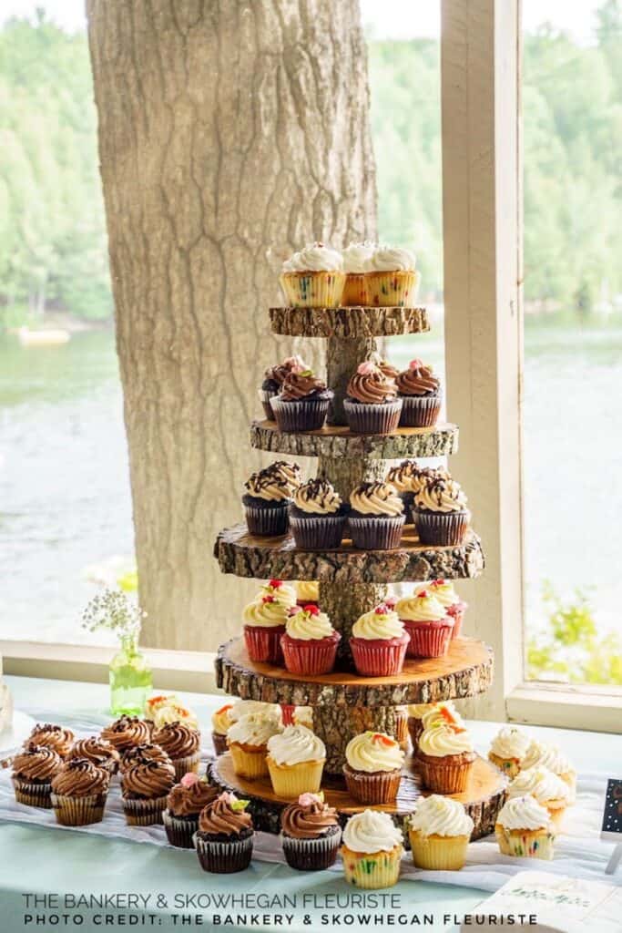 A tiered wooden cupcake stand displays an assortment of decorated cupcakes by a window with a lake view.