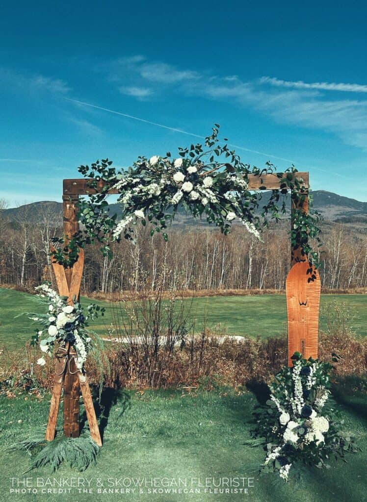 Wooden arch with white flowers, greenery, and ski-shaped accents on a grassy field, with mountains and trees in the background.