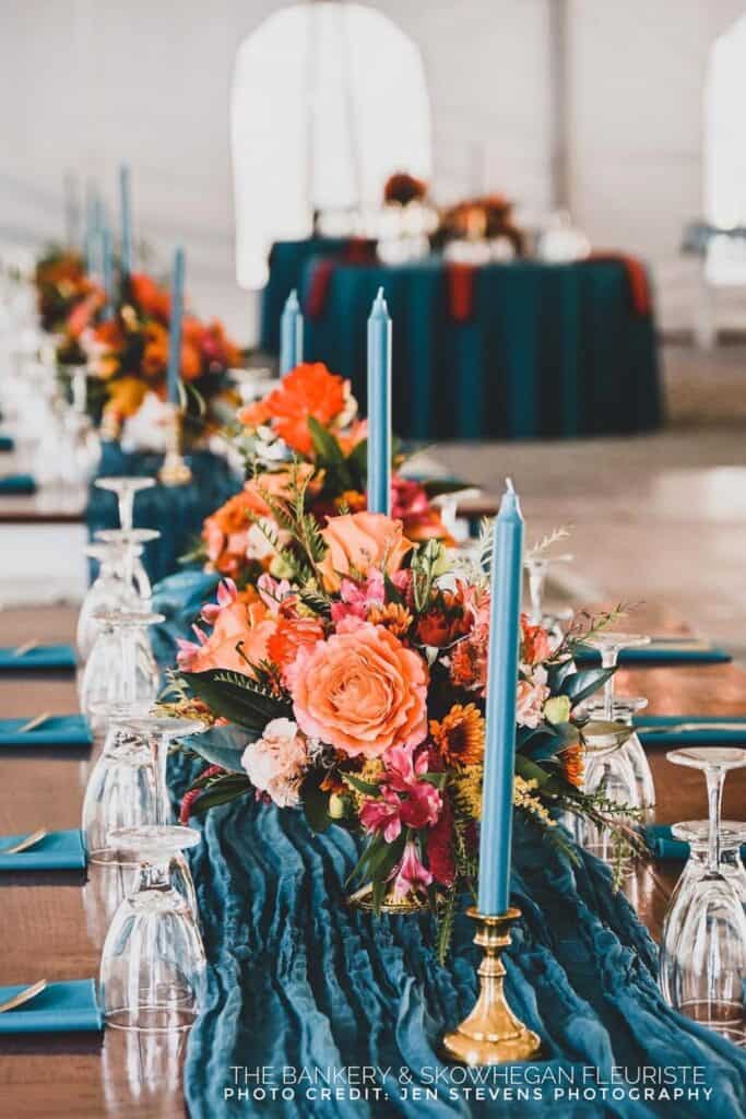 Elegant table setting with blue candles, orange and pink floral arrangement, and glassware on a textured blue runner.