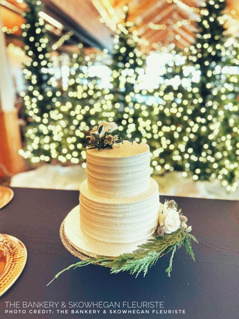 A white tiered cake with floral decorations sits on a table. In the background, Christmas trees are adorned with white lights.