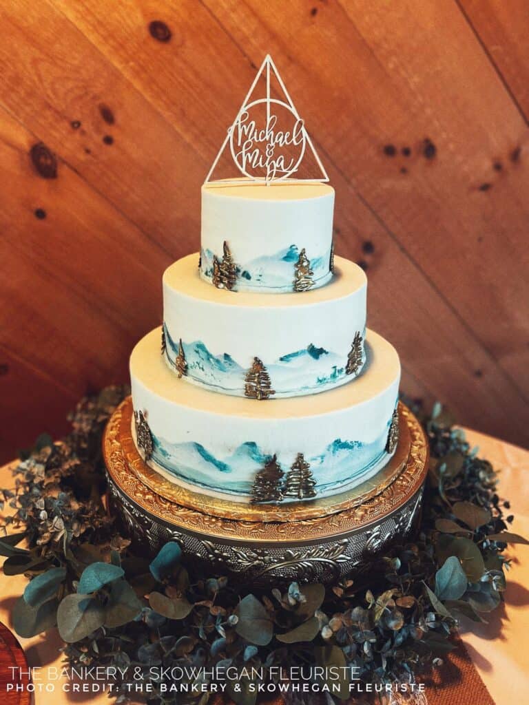 Three-tiered white cake with mountain scenery and trees, topped with a geometric decor piece on an ornate stand surrounded by greenery.