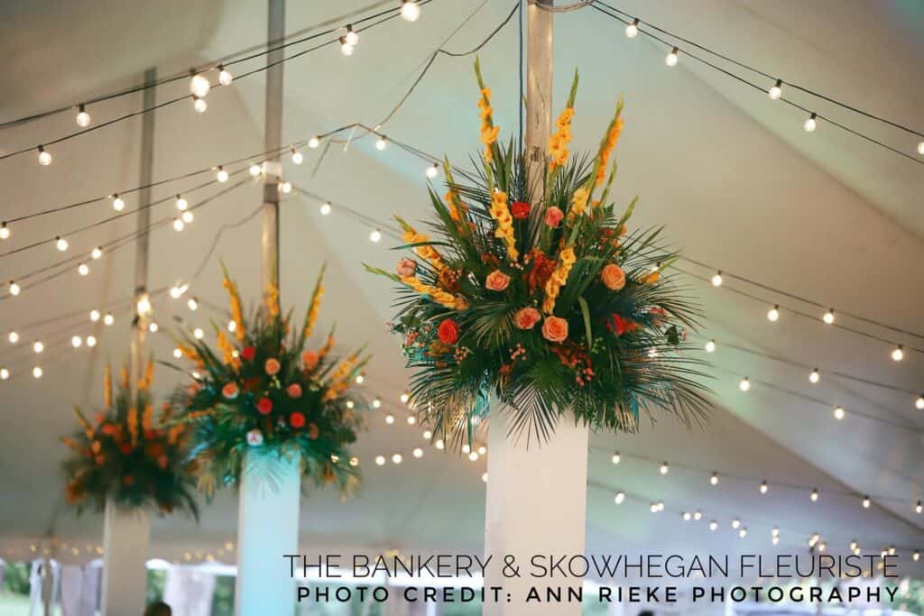 Floral arrangements with orange and yellow flowers hang from tent poles, surrounded by string lights.