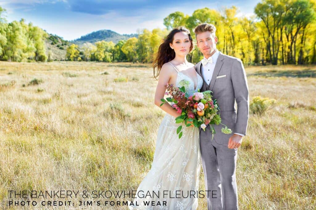 A bride in a white dress and a groom in a gray suit stand together holding a bouquet in an open, grassy field with trees in the background.