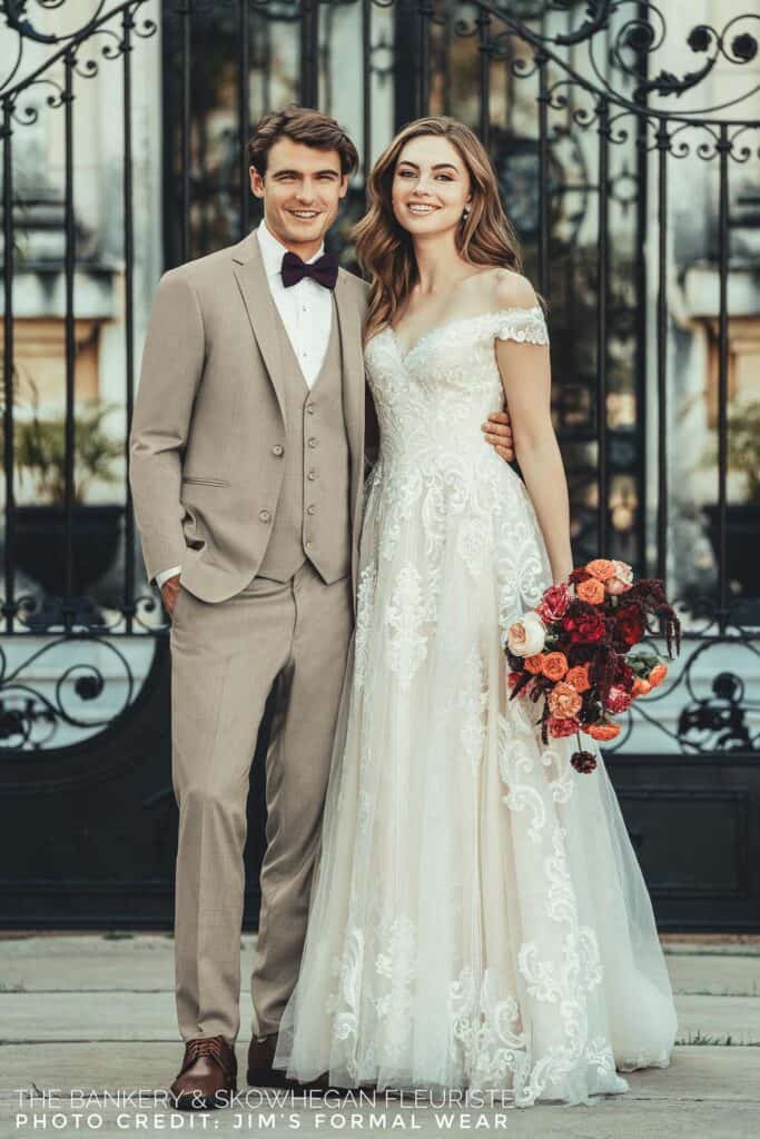 Bride in a white lace gown and groom in a gray suit smile by a decorative iron gate. She holds a bouquet of red and pink flowers.