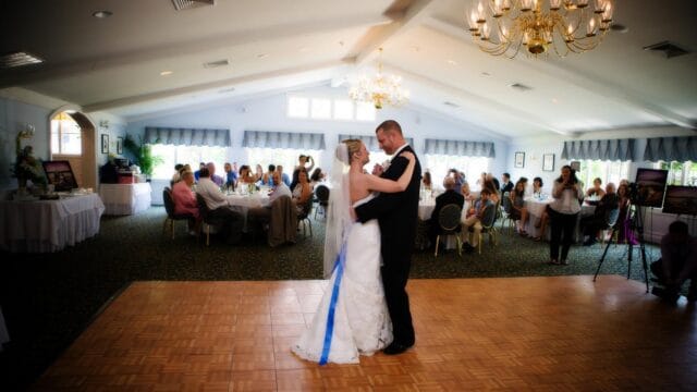 A bride and groom share a first dance on a wooden floor in a banquet hall, surrounded by seated guests at round tables, under decorative chandeliers.