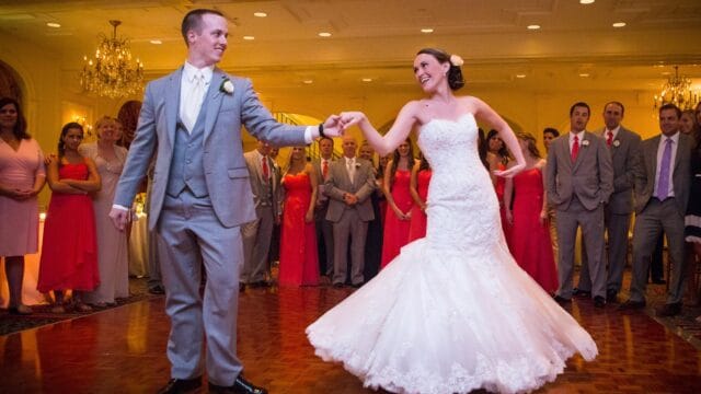 A bride and groom dance together on a ballroom floor, surrounded by guests in formal attire.