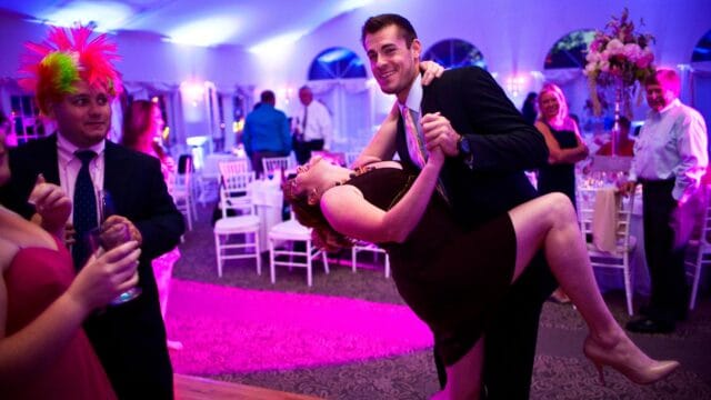 A couple dances enthusiastically at a reception under purple lighting. Nearby, a person with a colorful wig observes while holding a drink.