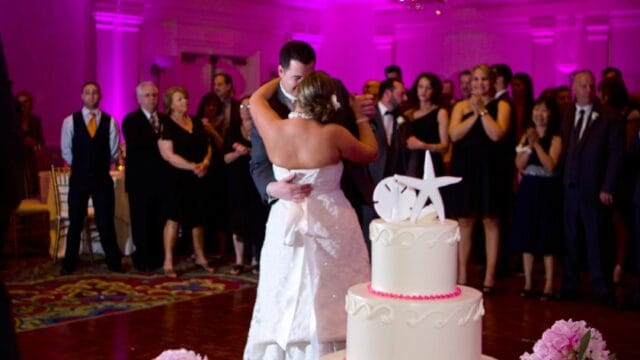 A couple embraces on the dance floor at a wedding reception, with a three-tiered cake featuring a starfish and pink flowers in the foreground. Guests watch in the background.