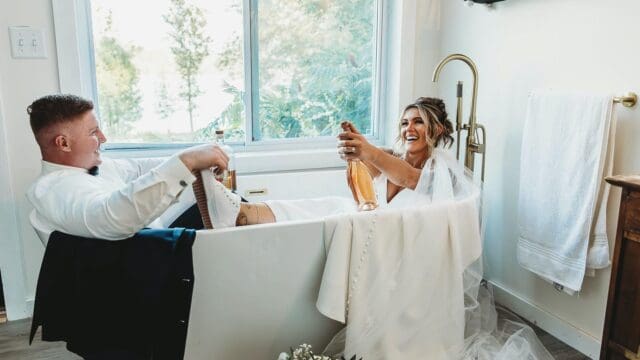 Bride and groom smile in a bathtub, holding champagne bottles. She wears a veil and dress; he wears a white shirt and bow tie, with a bright window behind them.