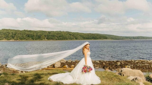 Bride in a white gown and long veil holds a colorful bouquet, standing by a rocky shoreline with trees and a cloudy sky in the background.