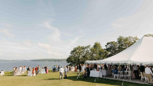 Outdoor gathering by a lake with people mingling near a large white tent.