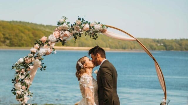 A couple kisses under a floral arch by a lakeside, with a grassy field and trees in the background.