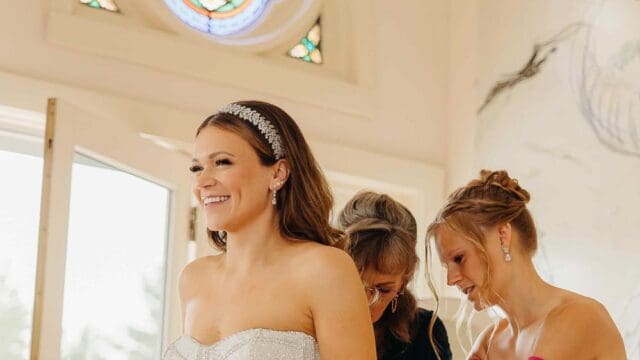 Bride in a strapless wedding gown stands smiling as two women assist her. A stained glass window is visible in the background.