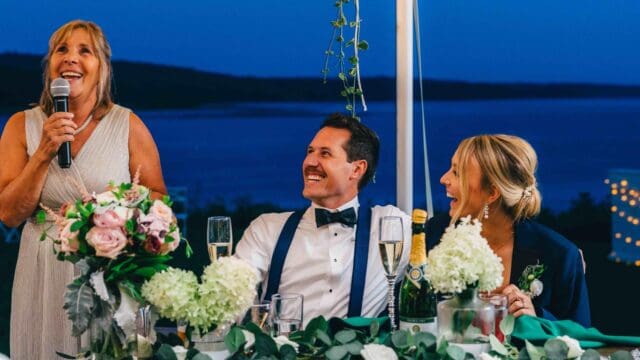 A woman speaks into a microphone at a wedding reception as a couple in formal attire smiles and listens at a flower-adorned table.