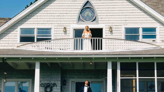 A bride stands on a balcony above a house, while a groom stands below on a porch, looking upward.