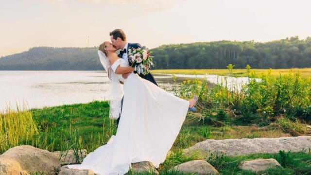 A bride in a white dress and veil holds a bouquet as the groom holds her leaning back, posing outdoors near a lake.