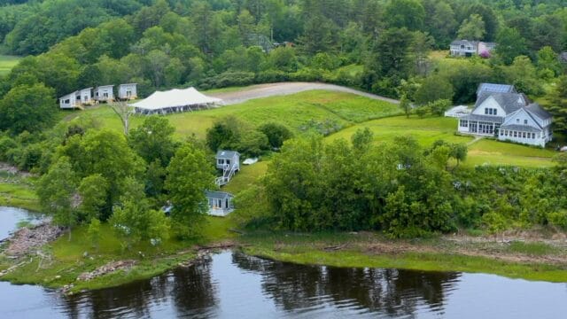 Aerial view of a riverside property with a large white tent, multiple buildings, and lush green surroundings.