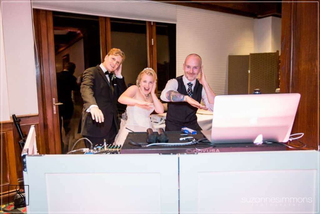 A bride and two men stand behind a DJ booth, smiling and posing while mimicking a fun gesture with one hand each.