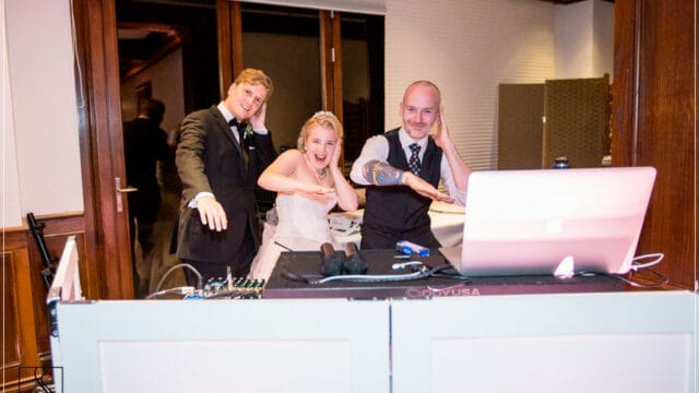 A bride and two men stand behind a DJ booth, smiling and posing while mimicking a fun gesture with one hand each.