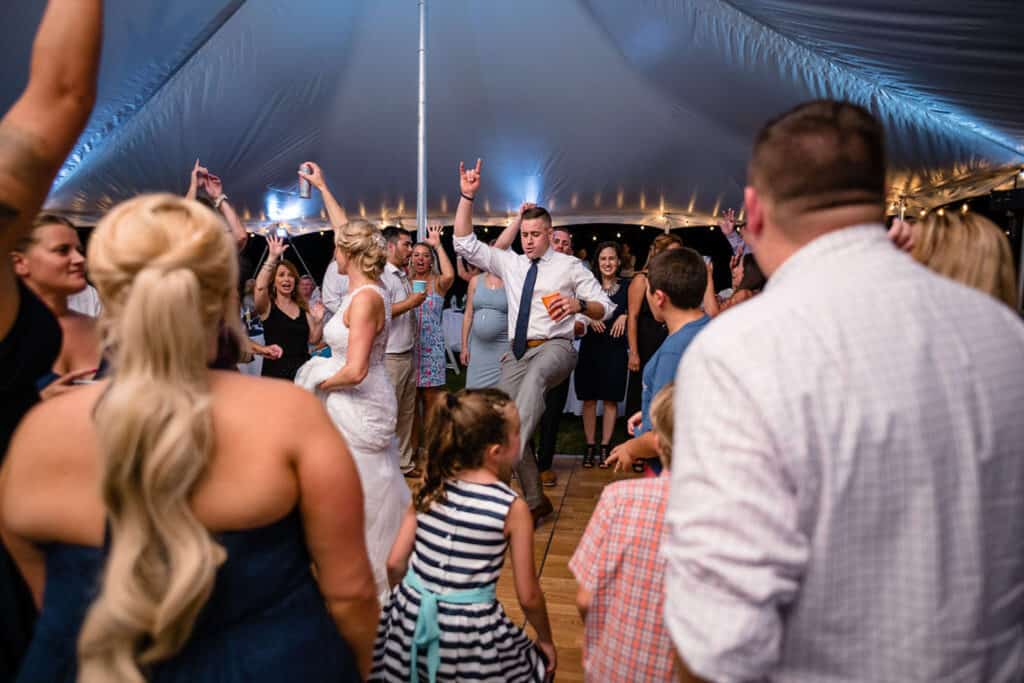 A wedding reception under a tent with people dancing. The bride in a white dress, and a man in a tie holds a drink while enthusiastically lifting a leg.