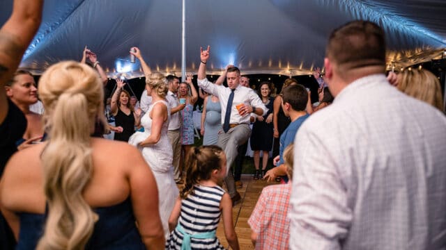 A wedding reception under a tent with people dancing. The bride in a white dress, and a man in a tie holds a drink while enthusiastically lifting a leg.