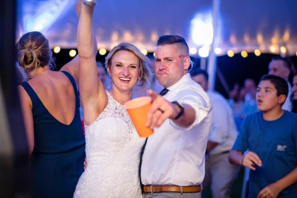 Bride and groom smiling and gesturing toward the camera at an outdoor evening event with string lights. Other people, including one in a blue shirt, are visible.