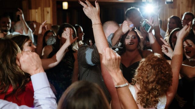 People dancing and celebrating indoors, with raised hands and visible excitement. A bright camera flash is visible in the background.