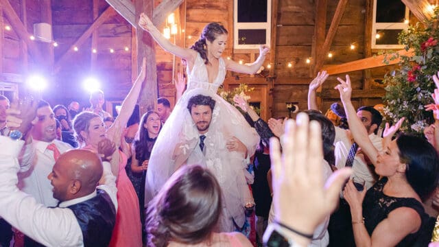 A joyful bride, lifted by guests, celebrates with her wedding party on the dance floor in a rustic indoor venue with string lights.