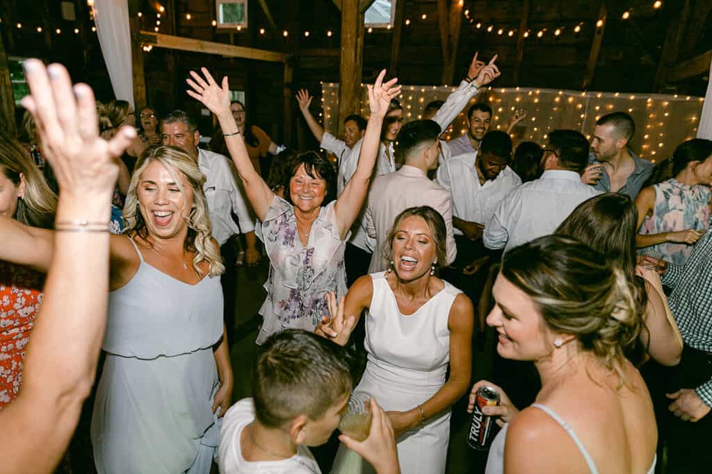 A group of men and women dancing and celebrating in a warmly lit venue. Some hold drinks, while others raise their hands, smile, and chat, enjoying the event.