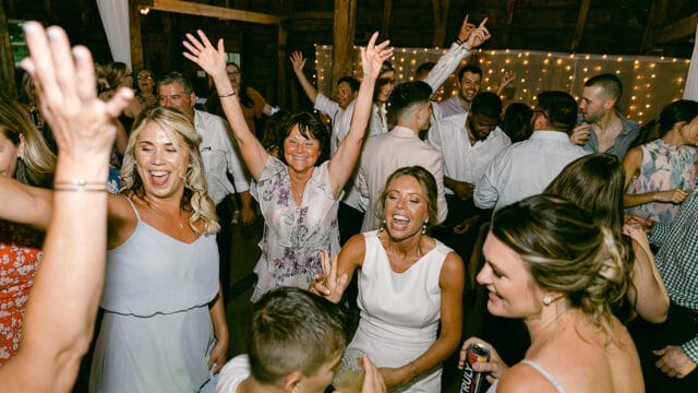 A group of men and women dancing and celebrating in a warmly lit venue. Some hold drinks, while others raise their hands, smile, and chat, enjoying the event.