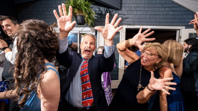 A group of people enthusiastically dancing at a social gathering, with a man in a suit and a woman in a black dress playfully raising their hands.