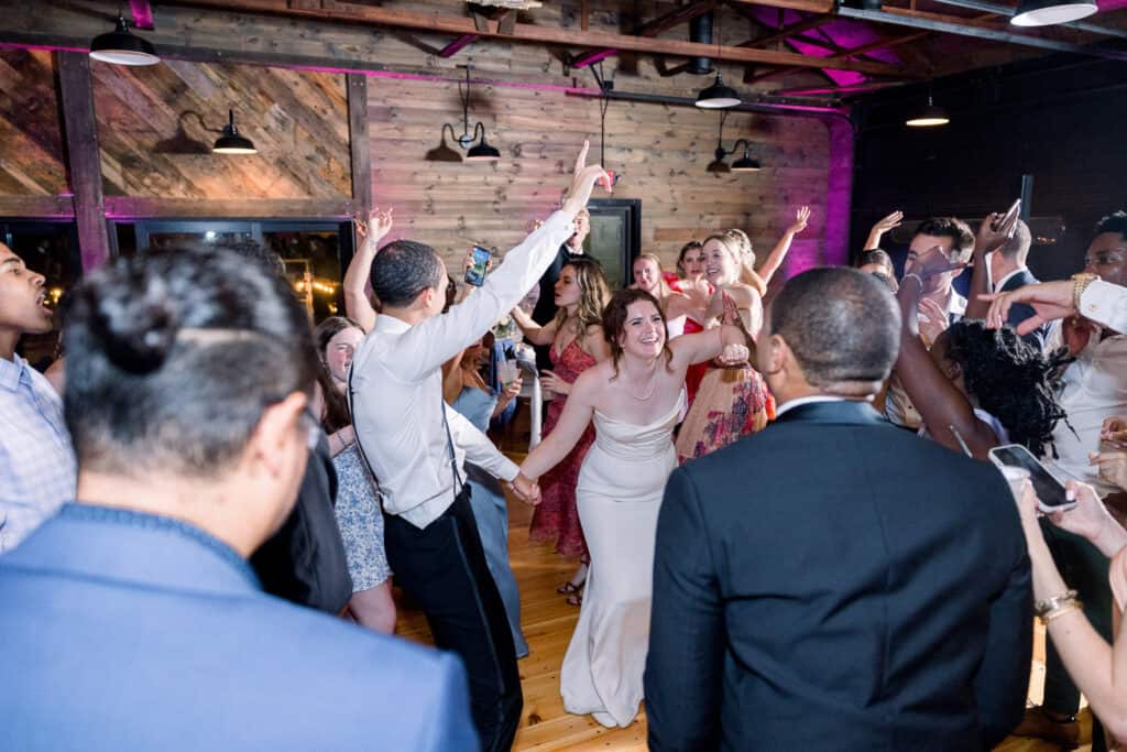 A group of people, including a bride in a white dress and a man in a white shirt, dance energetically at a wedding reception in a rustic, wood-paneled venue.