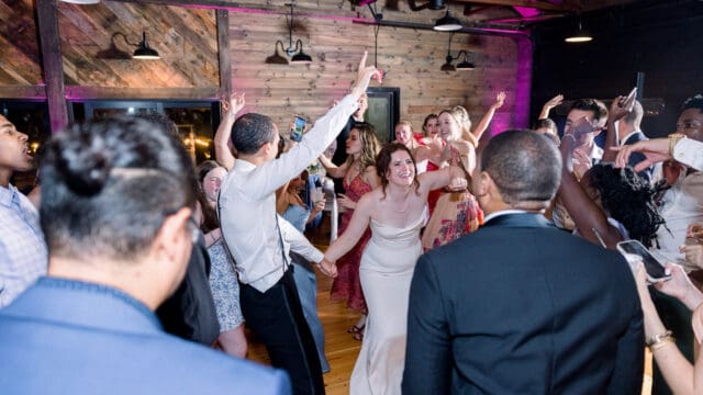 A group of people, including a bride in a white dress and a man in a white shirt, dance energetically at a wedding reception in a rustic, wood-paneled venue.