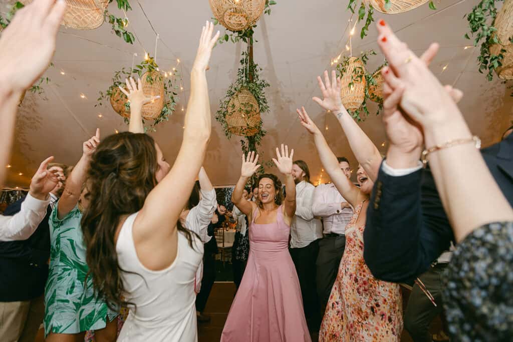 A group of people with raised hands, dancing together in a room with hanging lights and greenery. The woman in the center is smiling and wearing a pink dress.