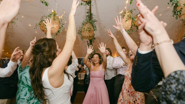 A group of people with raised hands, dancing together in a room with hanging lights and greenery. The woman in the center is smiling and wearing a pink dress.