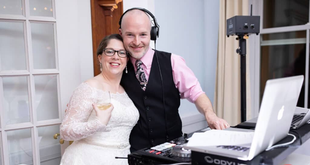 A smiling bride in a white dress stands next to a DJ wearing headphones. They are inside a well-lit room with sound equipment on a table.