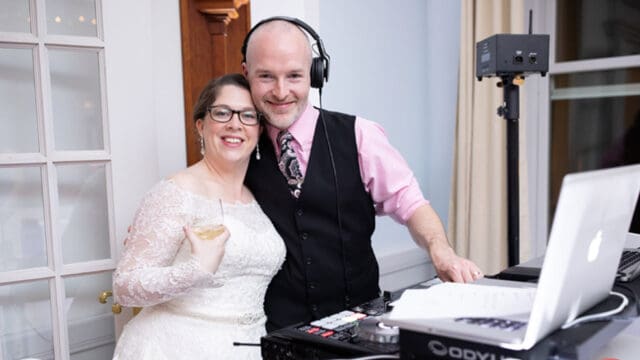 A smiling bride in a white dress stands next to a DJ wearing headphones. They are inside a well-lit room with sound equipment on a table.