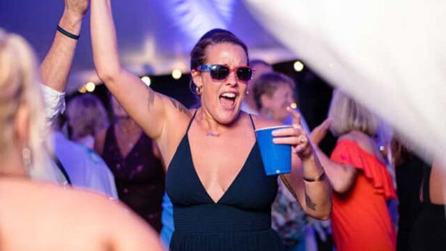 A woman wearing sunglasses holds a blue cup and raises her other hand while singing or shouting at a lively event with people around her.