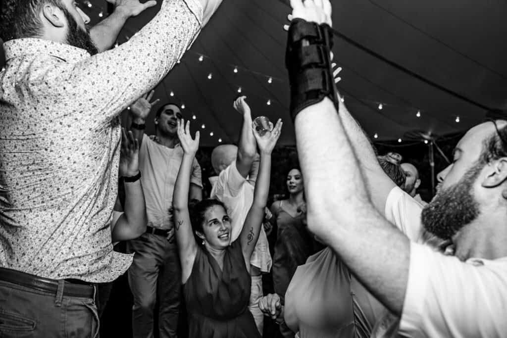 A group of people dance energetically with raised arms at a lively event under string lights; one person in the foreground has a cast on their arm.