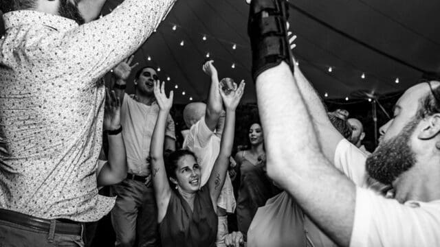 A group of people dance energetically with raised arms at a lively event under string lights; one person in the foreground has a cast on their arm.