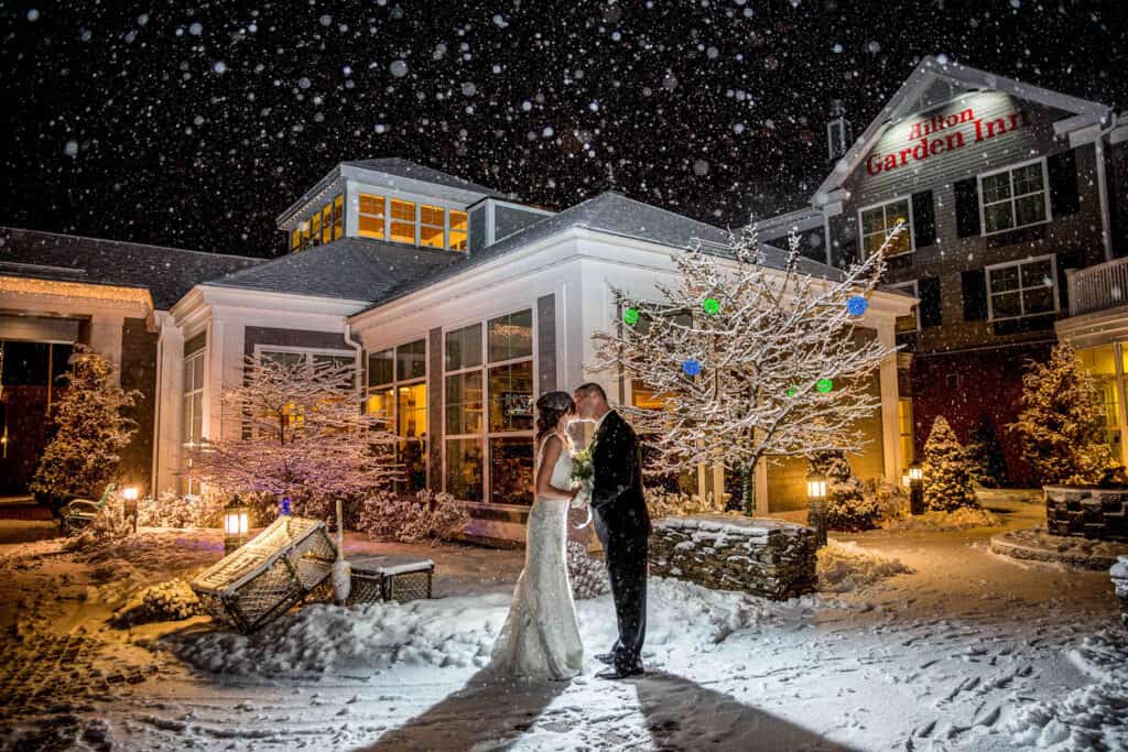 Bride and groom kiss in front of a Hilton Garden Inn at night during a snowstorm, surrounded by snow-covered trees and decor, with the lit hotel behind them.