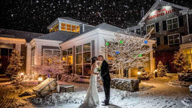 Bride and groom kiss in front of a Hilton Garden Inn at night during a snowstorm, surrounded by snow-covered trees and decor, with the lit hotel behind them.