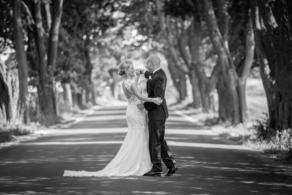 Bride in a lace dress with a train and groom in a suit embrace on a tree-lined road. The black-and-white scene highlights their intimate moment.