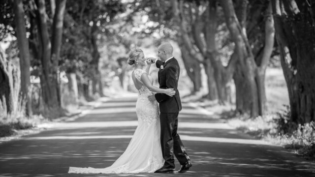 Bride in a lace dress with a train and groom in a suit embrace on a tree-lined road. The black-and-white scene highlights their intimate moment.